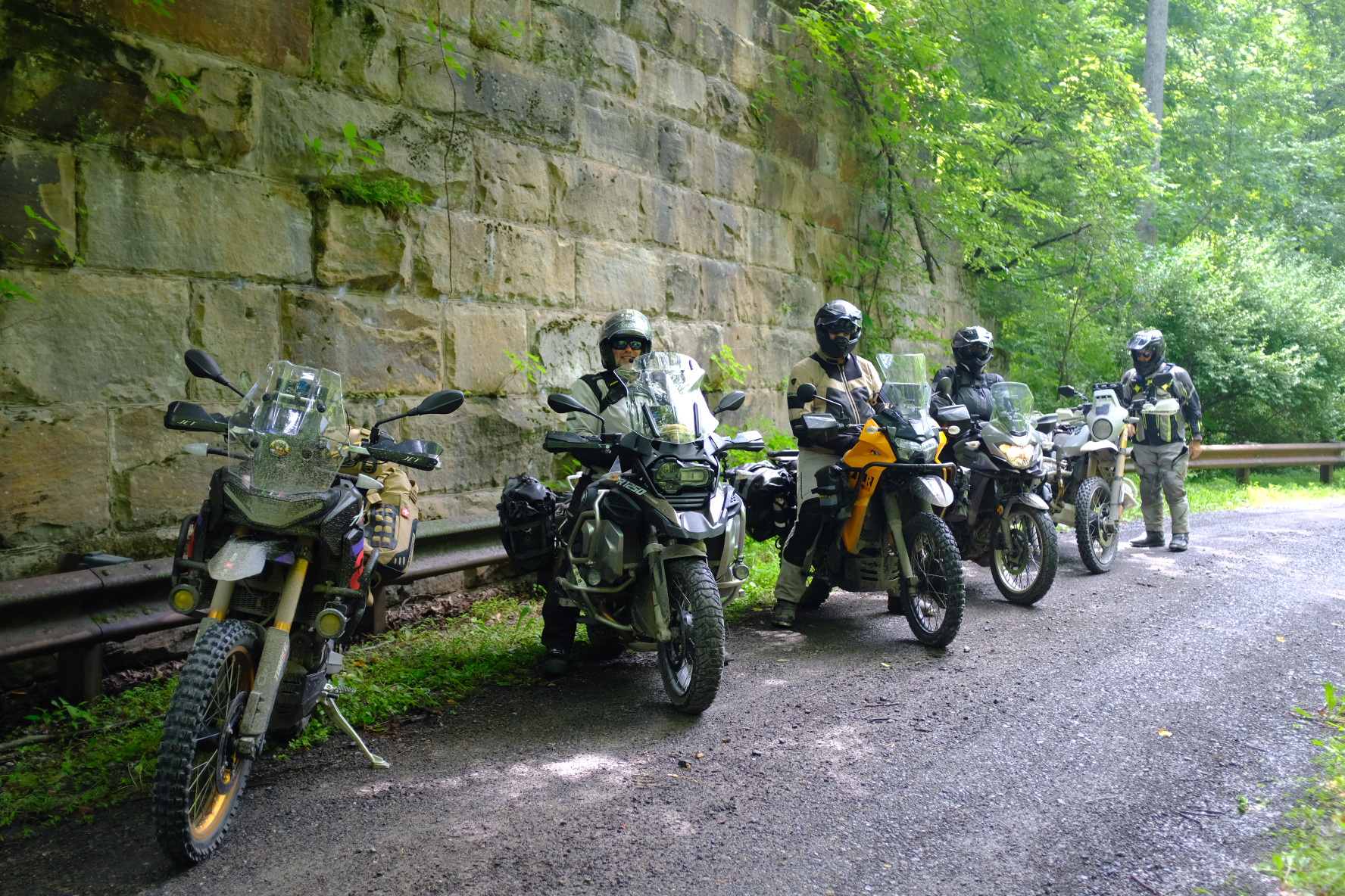 Adventure bikes lined up along a stone retaining wall on a shaded gravel road