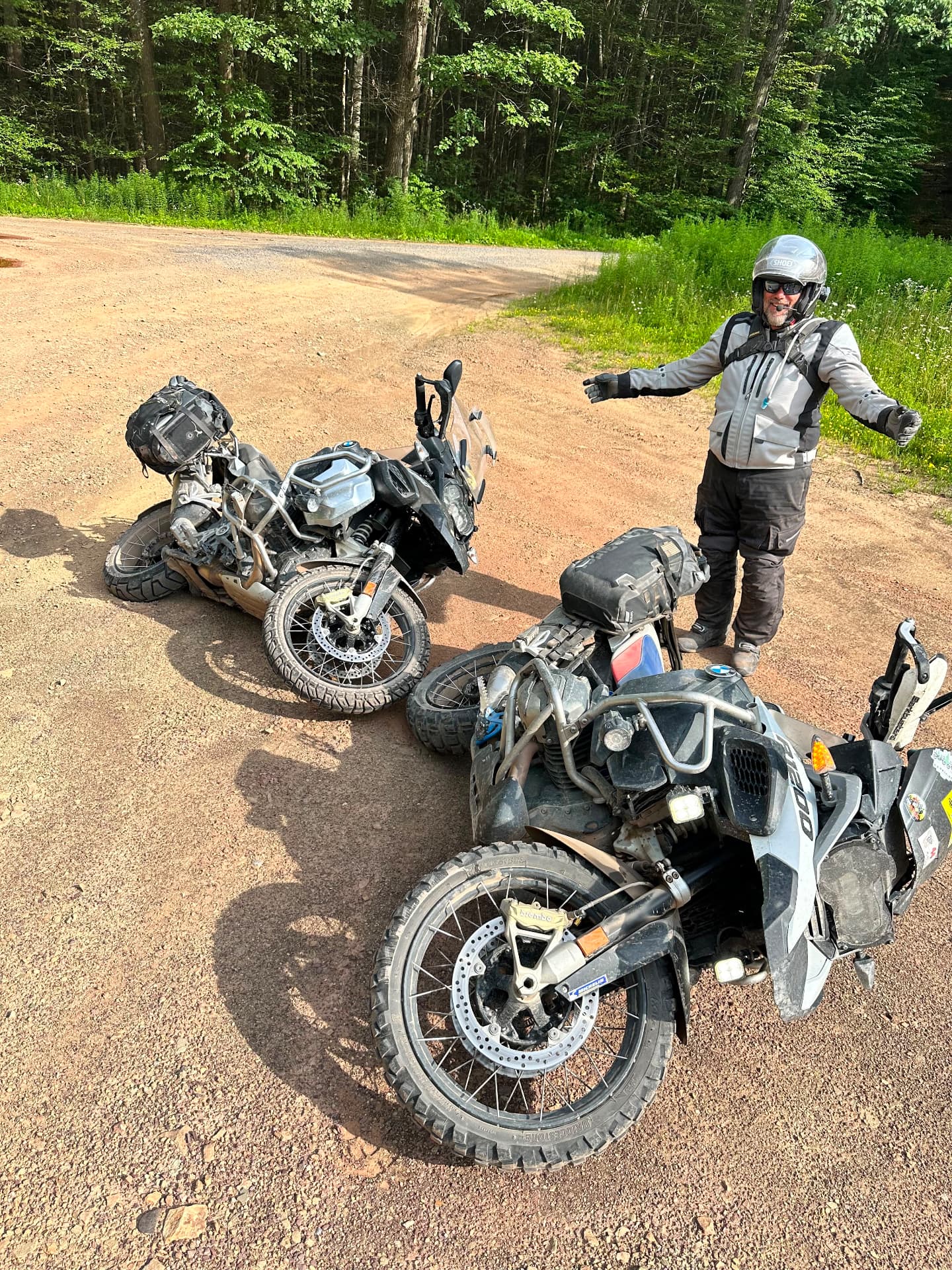 Rider shrugging beside two adventure bikes laid over on a dirt forest road